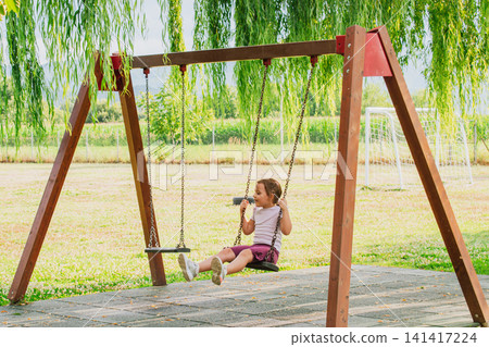 Side view of cute little girl swinging high on wooden swing set at outdoor park playground under green willow branches during warm sunny summer day Side view of cute little girl swinging high on wooden swing set at outdoor park playground under green willow branches during warm sunny summer day 141417224
