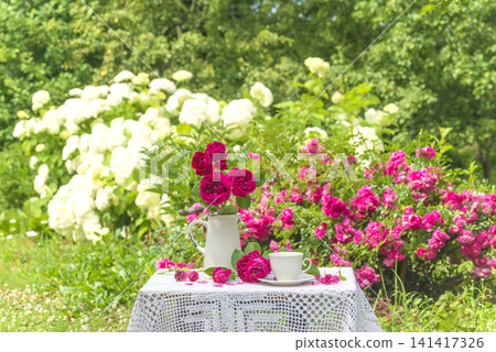Summer leisure activities in the garden; white cup of tea, bouquet of rose flowers on a table covered by lacy tablecloth in the summer blooming garden 141417326