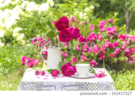Summer leisure activities in the garden; white cup of tea, bouquet of rose flowers on a table covered by lacy tablecloth in the summer blooming garden 141417327