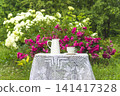 Summer leisure activities in the garden; white cup of tea, bouquet of rose flowers on a table covered by lacy tablecloth in the summer blooming garden 141417328
