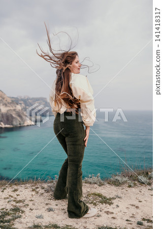 Woman cliff ocean with wind-blown hair stands on a rocky edge overlooking the blue sea during a scenic trip Woman cliff ocean with wind-blown hair stands on a rocky edge overlooking the blue sea during a scenic trip 141418317