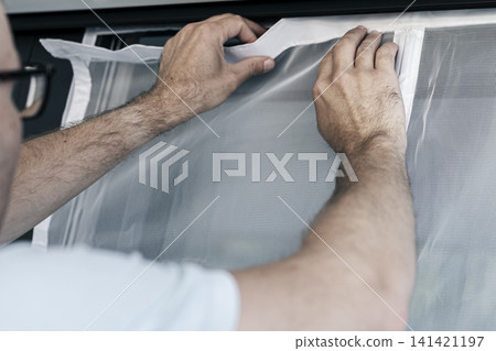 Person securing a white magnetic mosquito screen into the frame of balcony door, illustrating precise home task and maintenance. 141421197