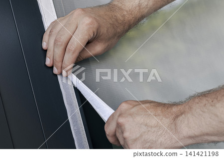 Hands securing a white mosquito screen into the window frame, illustrating precise handwork in home tasks and maintenance. Hands securing a white mosquito screen into the window frame, illustrating precise handwork in home tasks and maintenance. 141421198