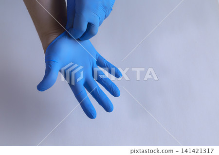 Hands pull blue medical gloves onto fingers against gray wall. 141421317