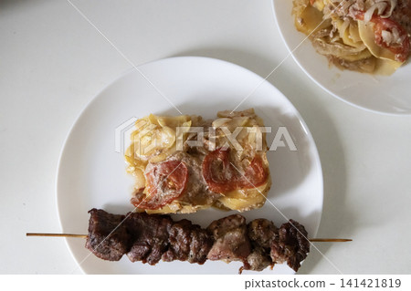 Delicious baked meat skewer served on a white plate with potato casserole, tomatoes and onions, ready for a tasty homemade lunch or dinner meal, top view close up. Delicious baked meat skewer served on a white plate with potato casserole, tomatoes and onions, ready for a tasty homemade lunch or dinner meal, top view close up. 141421819