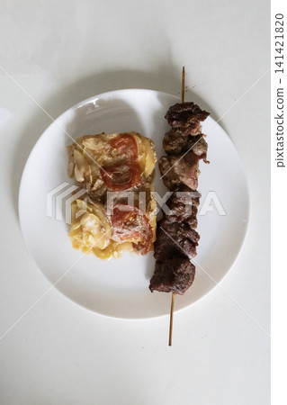 Delicious baked meat skewer served on a white plate with potato casserole, tomatoes and onions, ready for a tasty homemade lunch or dinner meal, top view close up. Delicious baked meat skewer served on a white plate with potato casserole, tomatoes and onions, ready for a tasty homemade lunch or dinner meal, top view close up. 141421820