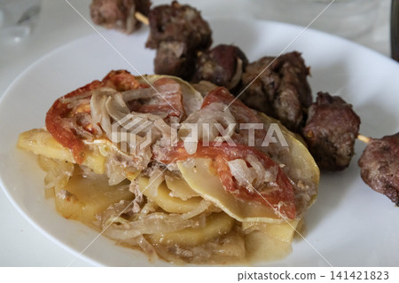 Delicious baked meat skewer served on a white plate with potato casserole, tomatoes and onions, ready for a tasty homemade lunch or dinner meal, top view close up. Delicious baked meat skewer served on a white plate with potato casserole, tomatoes and onions, ready for a tasty homemade lunch or dinner meal, top view close up. 141421823