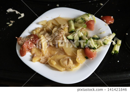 Leftover food on a white plate: remains of baked potato casserole with tomatoes and a fresh cucumber and cabbage salad on a dark background. 141421846