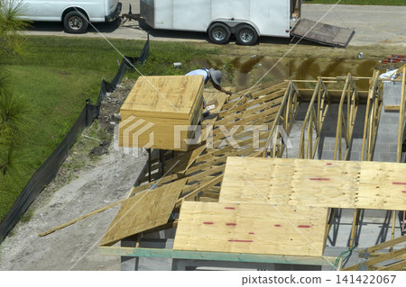 Builders working on roof construction of unfinished residential house with wooden frame structure in Florida suburban area. Housing development concept Builders working on roof construction of unfinished residential house with wooden frame structure in Florida suburban area. Housing development concept 141422067