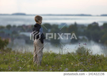 Back of young child boy alone on grassy hill top on foggy village among green trees background. Back of young child boy alone on grassy hill top on foggy village among green trees background. 141422136
