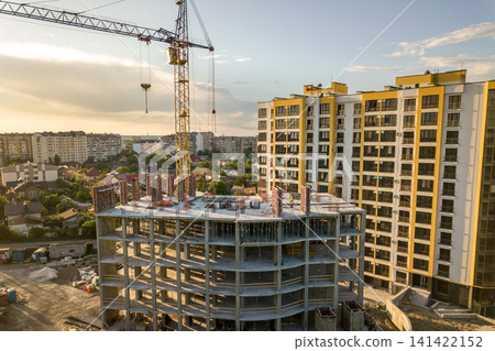 Apartment or office tall building under construction. Brick walls, glass windows, scaffolding and concrete support pillars. Tower crane on bright blue sky copy space background. Apartment or office tall building under construction. Brick walls, glass windows, scaffolding and concrete support pillars. Tower crane on bright blue sky copy space background. 141422152