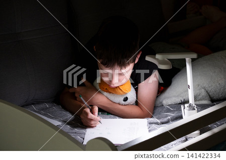 Schoolboy doing homework in bed under desk lamp in the evening. Child writing in notebook at home, education, learning, concentration and cozy bedroom atmosphere. 141422334