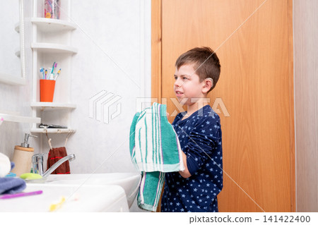 A boy wearing hearing aids and pajamas wipes his face with a towel in front of a mirror above the sink. A child with hearing aids performs household chores A boy wearing hearing aids and pajamas wipes his face with a towel in front of a mirror above the sink. A child with hearing aids performs household chores 141422400