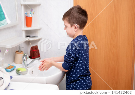 A boy wearing hearing aids and pajamas washes his hands with water in front of a mirror above the sink. A child with hearing aids performs household chores A boy wearing hearing aids and pajamas washes his hands with water in front of a mirror above the sink. A child with hearing aids performs household chores 141422402