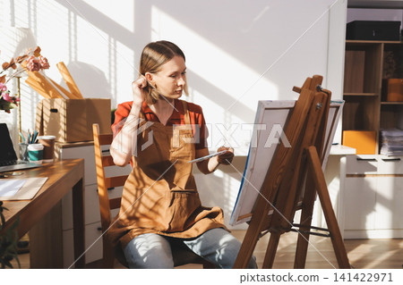 Picture of serious concentrated young Caucasian female artist sitting at desk with painting accessories, holding tube of oil paint, mixing colors on palette; unfinished painting on canvas near her 141422971
