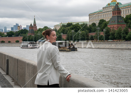 Woman turns toward camera while walking along Sofiyskaya Embankment in Moscow with Kremlin and Moscow River behind. Urban lifestyle, sightseeing, travel, architecture, confidence, city exploration Woman turns toward camera while walking along Sofiyskaya Embankment in Moscow with Kremlin and Moscow River behind. Urban lifestyle, sightseeing, travel, architecture, confidence, city exploration 141423005
