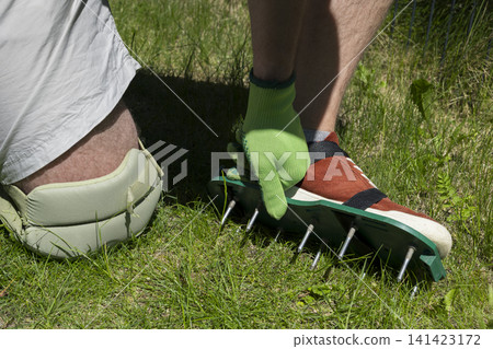 Person wearing aerator shoes on a lawn, illustrating home gardening tools and simple manual soil care for seasonal yard work. Person wearing aerator shoes on a lawn, illustrating home gardening tools and simple manual soil care for seasonal yard work. 141423172