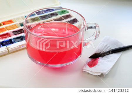 Workspace of a watercolor artist with glass of water, a brush, a stained napkin, and a palette with watercolor paints on a white table. Workspace of a watercolor artist with glass of water, a brush, a stained napkin, and a palette with watercolor paints on a white table. 141423212