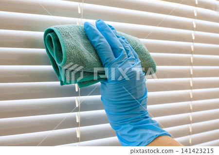 Young woman's hand wipes the blinds with a turquoise cloth. Rubber blue gloved hand holds a dirty cleaning cloth. Cleaning service concept 141423215
