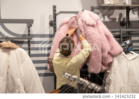 Woman shopper choosing trendy winter jacket in clothing store. Customer looking at price tag on fluffy pink coat in boutique. Lifestyle retail therapy and fashion consumerism concept. 141423232