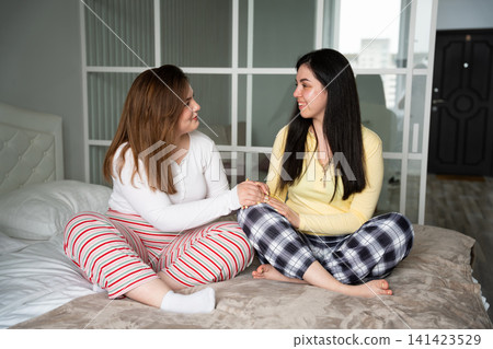 Two sisters sitting on bed having heart to heart conversation in cozy bedroom. Family bonding emotional support and sisterhood Two sisters sitting on bed having heart to heart conversation in cozy bedroom. Family bonding emotional support and sisterhood 141423529