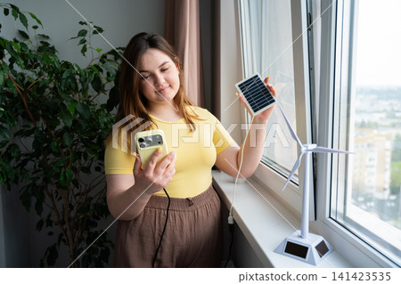 Young woman using smartphone and holding solar panel near window with model wind turbine. Renewable energy technology and smart energy monitoring app Young woman using smartphone and holding solar panel near window with model wind turbine. Renewable energy technology and smart energy monitoring app 141423535
