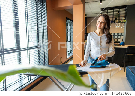 Long-haired young woman standing near the ironing board and ironing laundry Long-haired young woman standing near the ironing board and ironing laundry 141423603