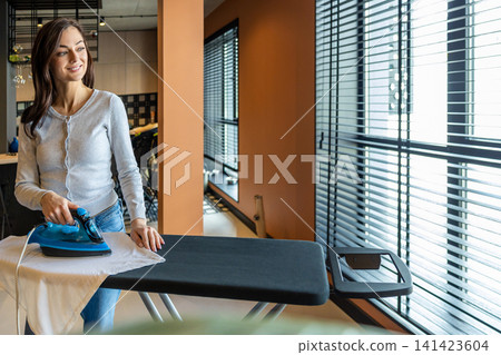 Long-haired young woman standing near the ironing board and ironing laundry Long-haired young woman standing near the ironing board and ironing laundry 141423604