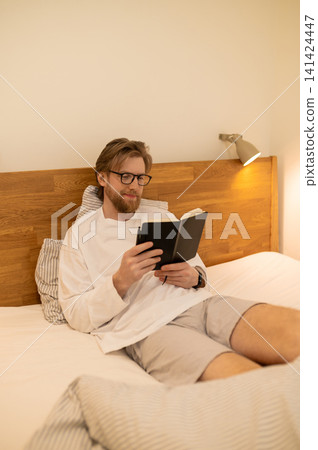 Fair-haired young man sitting in bed with a book in hands Fair-haired young man sitting in bed with a book in hands 141424447