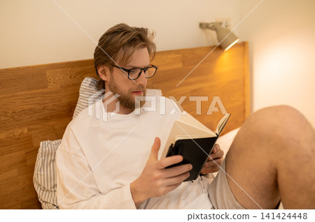 Fair-haired young man sitting in bed with a book in hands Fair-haired young man sitting in bed with a book in hands 141424448