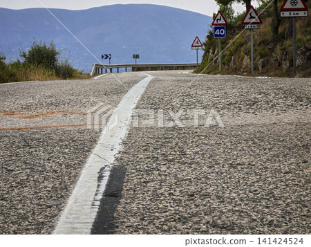 The view shows a winding road with asphalt surface leading to the Puerto de Beniarres mountain pass. Road signs indicate directions and speed limits. Surrounding hills and trees are visible. The view shows a winding road with asphalt surface leading to the Puerto de Beniarres mountain pass. Road signs indicate directions and speed limits. Surrounding hills and trees are visible. 141424524