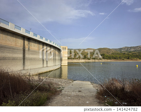 The large concrete arch dam wall of the Beniarres reservoir stretches along the blue water. The area shows land and vegetation near the shoreline. The large concrete arch dam wall of the Beniarres reservoir stretches along the blue water. The area shows land and vegetation near the shoreline. 141424782