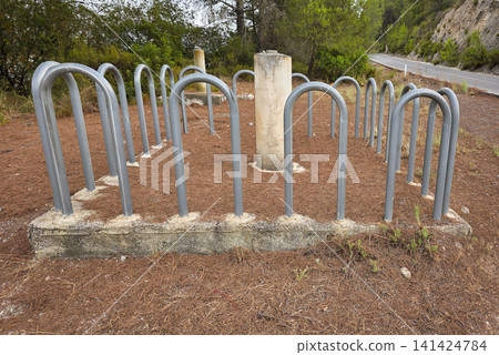 Bicycle parking racks made of metal are placed in a rest area covered with pine needles. The location is near a road by a reservoir. It is a typical outdoor area for cyclists. 141424784