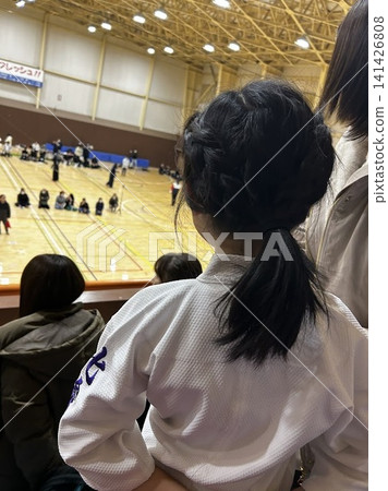 A girl in a martial arts uniform watches a martial arts match from the spectator seats in a gymnasium - Children's extracurricular activities and competitions A girl in a martial arts uniform watches a martial arts match from the spectator seats in a gymnasium - Children's extracurricular activities and competitions 141426808