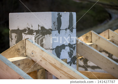 Close-up detail of roof frame of rough wooden lumber beams and chimney made of foam insulation blocks on blurred green background. Building, roofing, construction and renovation concept. Close-up detail of roof frame of rough wooden lumber beams and chimney made of foam insulation blocks on blurred green background. Building, roofing, construction and renovation concept. 141427787