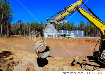 An excavator is lifting concrete drainage pipe at construction site surrounded by trees An excavator is lifting concrete drainage pipe at construction site surrounded by trees 141428477