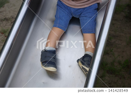 公園裡,一個穿著短褲的孩子正從溜滑梯上滑下來,露出他的腳。 公園裡,一個穿著短褲的孩子正從溜滑梯上滑下來,露出他的腳。 141428549