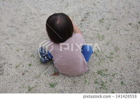 The back view of a child sitting on the ground in a park, playing in the sand. 141428558