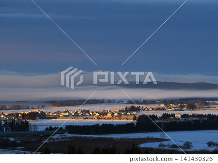 On a winter morning shrouded in mist, the rising sun shines through a gap in the fog. The snow-covered townscape emerges, appearing as if it were above the clouds, creating a striking and memorable scene. On a winter morning shrouded in mist, the rising sun shines through a gap in the fog. The snow-covered townscape emerges, appearing as if it were above the clouds, creating a striking and memorable scene. 141430326
