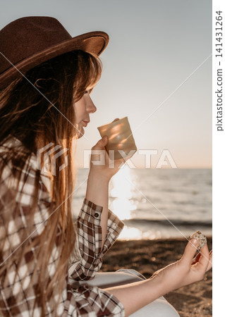 Woman hat sunset drinking from glass while sitting on beach during golden hour Woman hat sunset drinking from glass while sitting on beach during golden hour 141431264