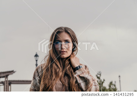 Woman portrait rain young woman wearing glasses and plaid jacket stands outdoors under an overcast sky with copy space Woman portrait rain young woman wearing glasses and plaid jacket stands outdoors under an overcast sky with copy space 141431295