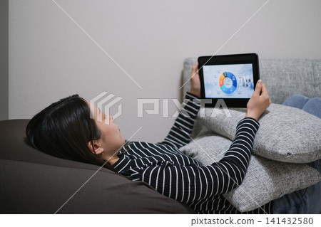 A woman is lying down and looking at a website about asset management on her tablet. A woman is lying down and looking at a website about asset management on her tablet. 141432580