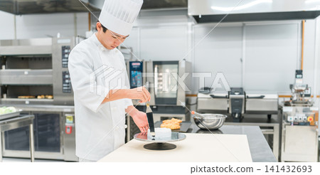 A young Asian man (pastry chef) in a chef's coat working in a confectionery workshop, cake shop kitchen, or bakery. A young Asian man (pastry chef) in a chef's coat working in a confectionery workshop, cake shop kitchen, or bakery. 141432693