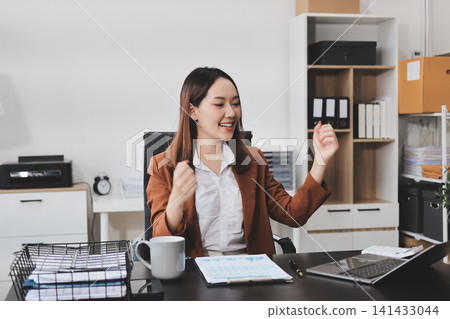 Young Asian professional business woman calculating finances and analyzing data charts on computer, smiling while working on accounting project in a modern office, planning budget Young Asian professional business woman calculating finances and analyzing data charts on computer, smiling while working on accounting project in a modern office, planning budget 141433044