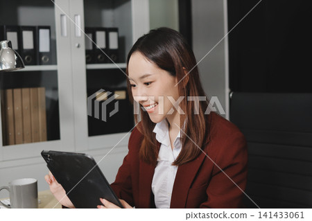 Smiling asian businesswoman talking on phone and using laptop while working at office desk Smiling asian businesswoman talking on phone and using laptop while working at office desk 141433061