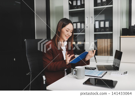 Smiling asian businesswoman talking on phone and using laptop while working at office desk 141433064