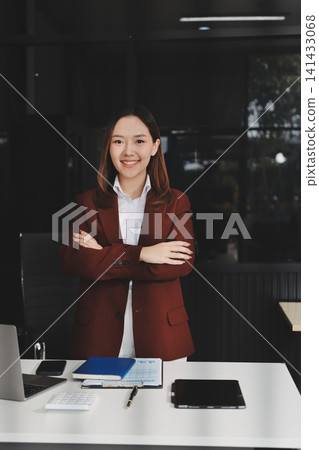 Smiling asian businesswoman talking on phone and using laptop while working at office desk Smiling asian businesswoman talking on phone and using laptop while working at office desk 141433068
