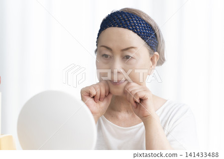 A senior woman uses a facial steamer to perform a self-care facial. 141433488