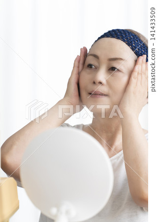 A senior woman uses a facial steamer to perform a self-care facial. 141433509