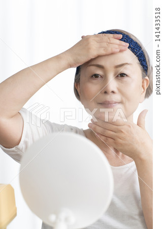 A senior woman uses a facial steamer to perform a self-care facial. 141433518